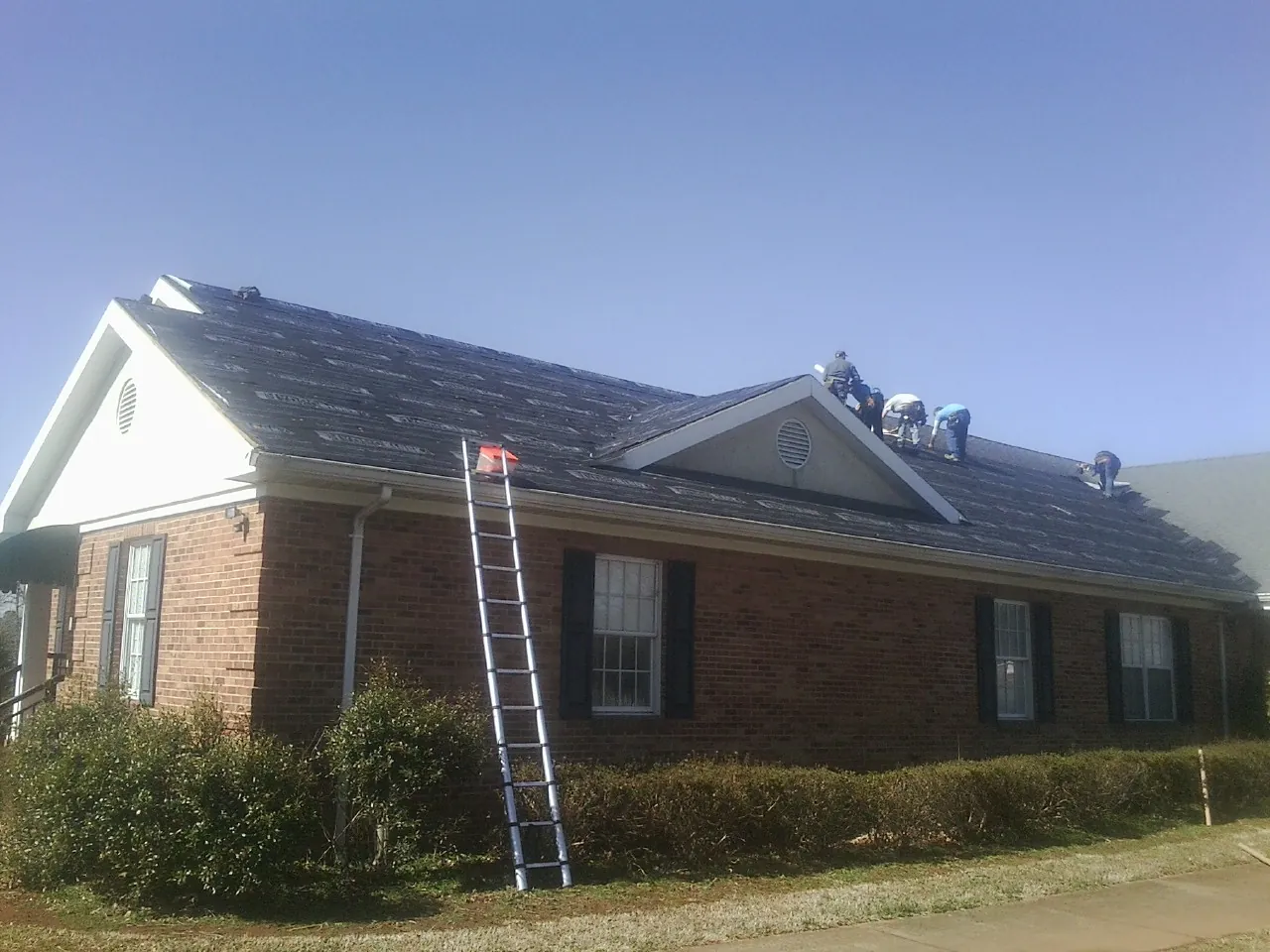 Crew tearing shingles off a brick ranch roof with a ladder set — Metal Roof Repair work by our crew in Hookerton
