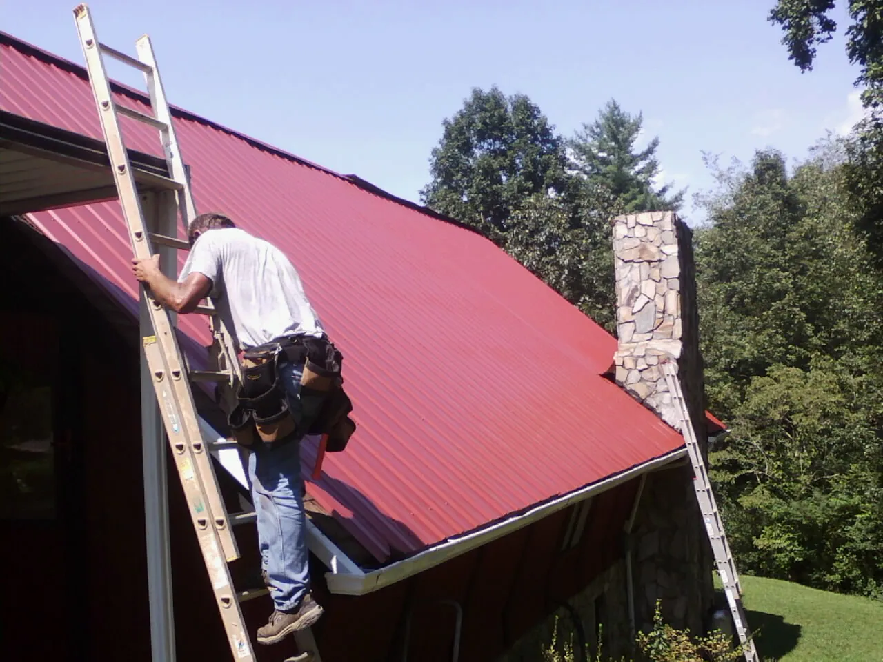 Roofer on a ladder beside a red metal roof and stone chimney — Soffit & Fascia Repair by Roofing Brothers in Hookerton